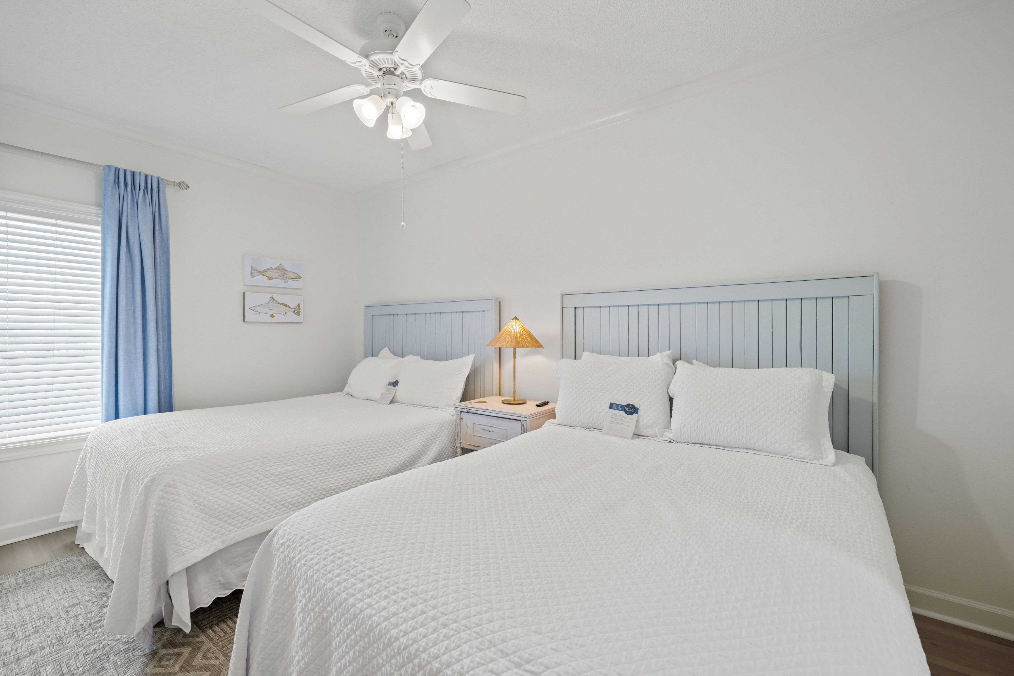 A hotel room with two beds, a ceiling fan, a nightstand with a lamp, and a window with blinds.