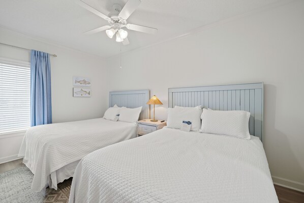 A hotel room with two beds, a ceiling fan, a nightstand with a lamp, and a window with blinds.