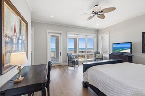 A hotel room with a bed, a desk, a chair, a TV, a ceiling fan, and a balcony with a view of the ocean.