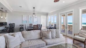 A spacious living area with a large sofa, dining table, and chairs, a kitchen area, and a balcony with a view of the beach. - Finntastic Beachfront House by Liquid Life (Gulf Shores)