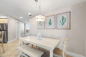 Un coin repas avec une table et un banc aux couleurs claires, un luminaire suspendu et trois reproductions encadrées de motifs botaniques accrochées au mur.