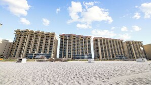 A beachfront hotel with multiple high-rise buildings, a sandy beach in the foreground, and a clear blue sky with clouds.