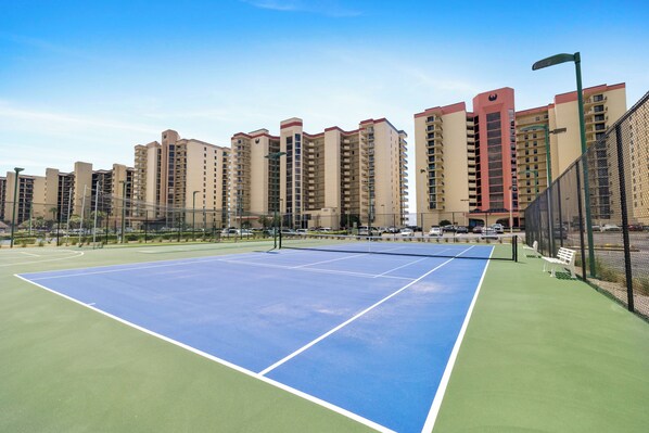 Een tennisbaan met een net, omringd door een hek van gaas, grenzend aan een wooncomplex.