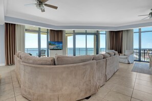 A spacious living room with a large beige sectional sofa, a flat-screen TV, and a balcony with a view of the ocean.