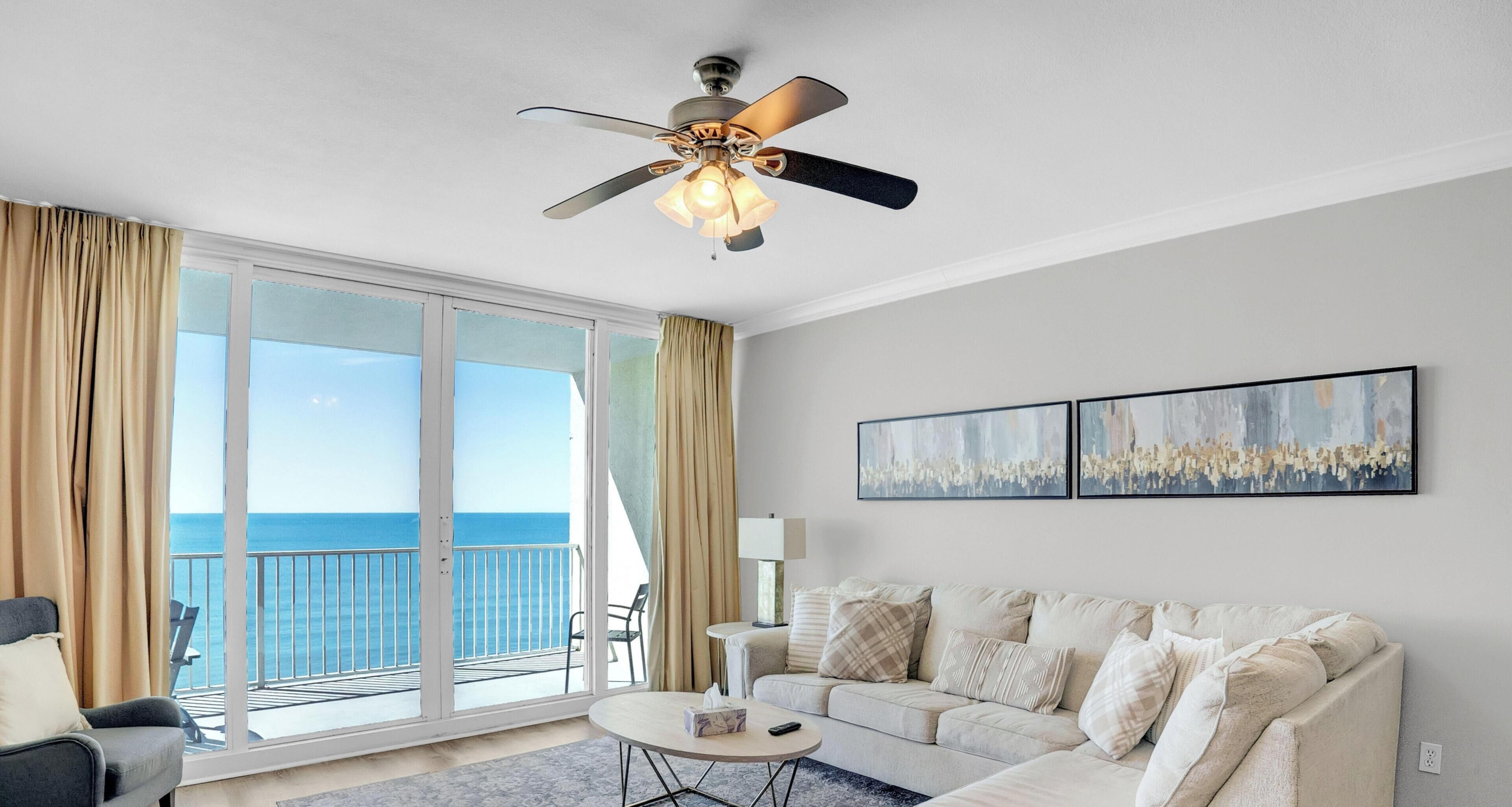 A hotel room with a balcony, a ceiling fan, a sofa, a coffee table, and two wall-mounted paintings.