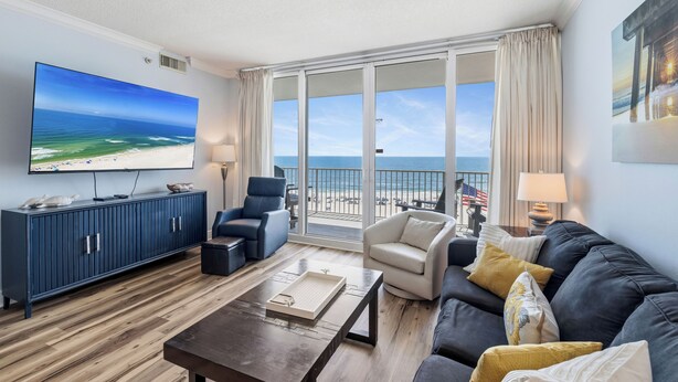 Una sala de estar con un televisor grande, un mueble azul, un sofá oscuro, un sillón blanco y vista a la playa.