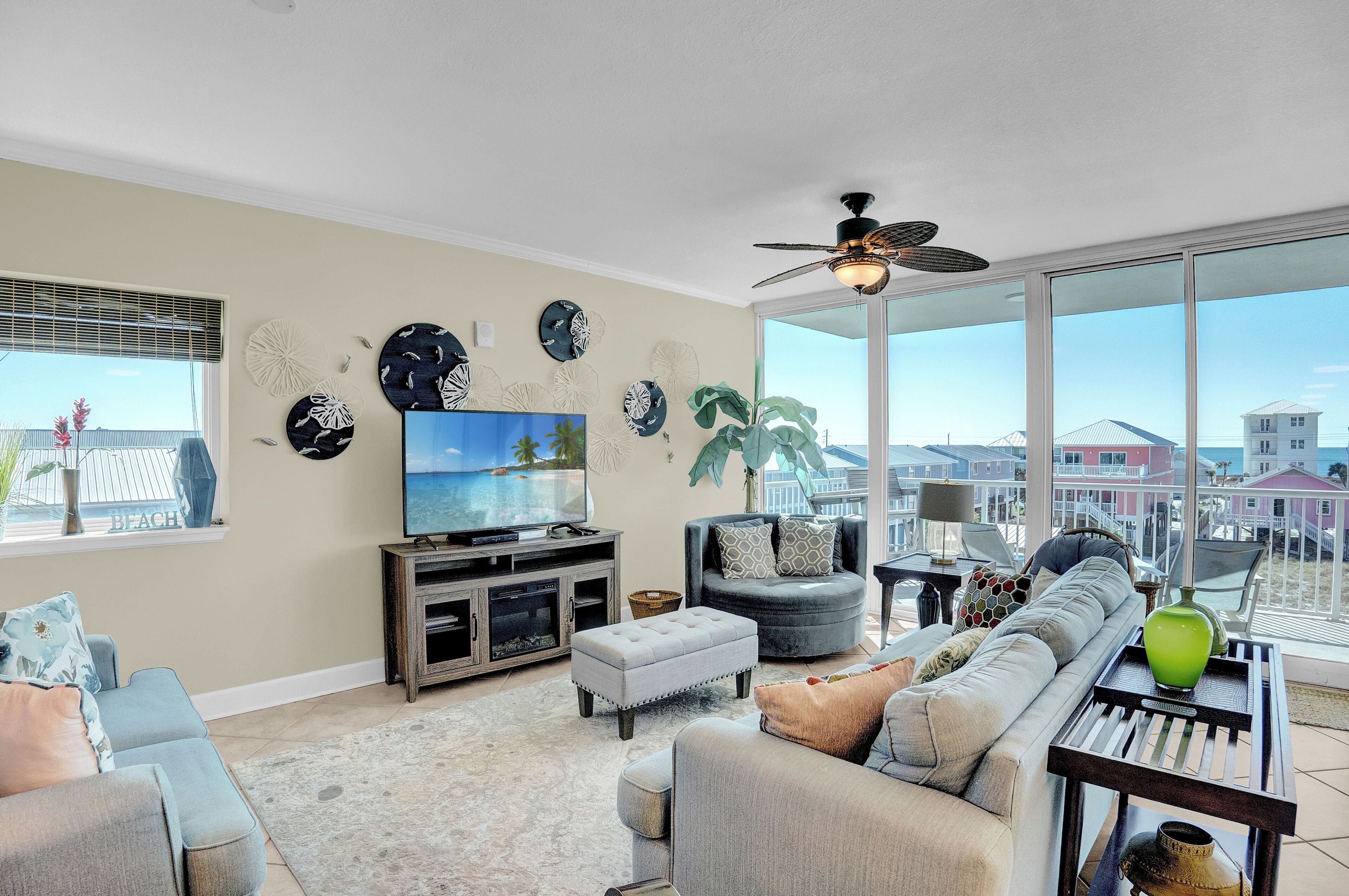 A spacious living room with a large TV, comfortable seating, a ceiling fan, and a balcony with a view.