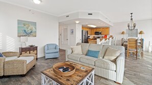Ein geräumiges Wohnzimmer mit einem großen, gestreiften Sofa, einem Korbsessel und einem blauen Sessel. Ein Couchtisch aus Holz mit einem Korb und Fernbedienungen steht in der Mitte. Im Hintergrund ist eine Küche mit Holzschränken und einem Essbereich zu sehen.