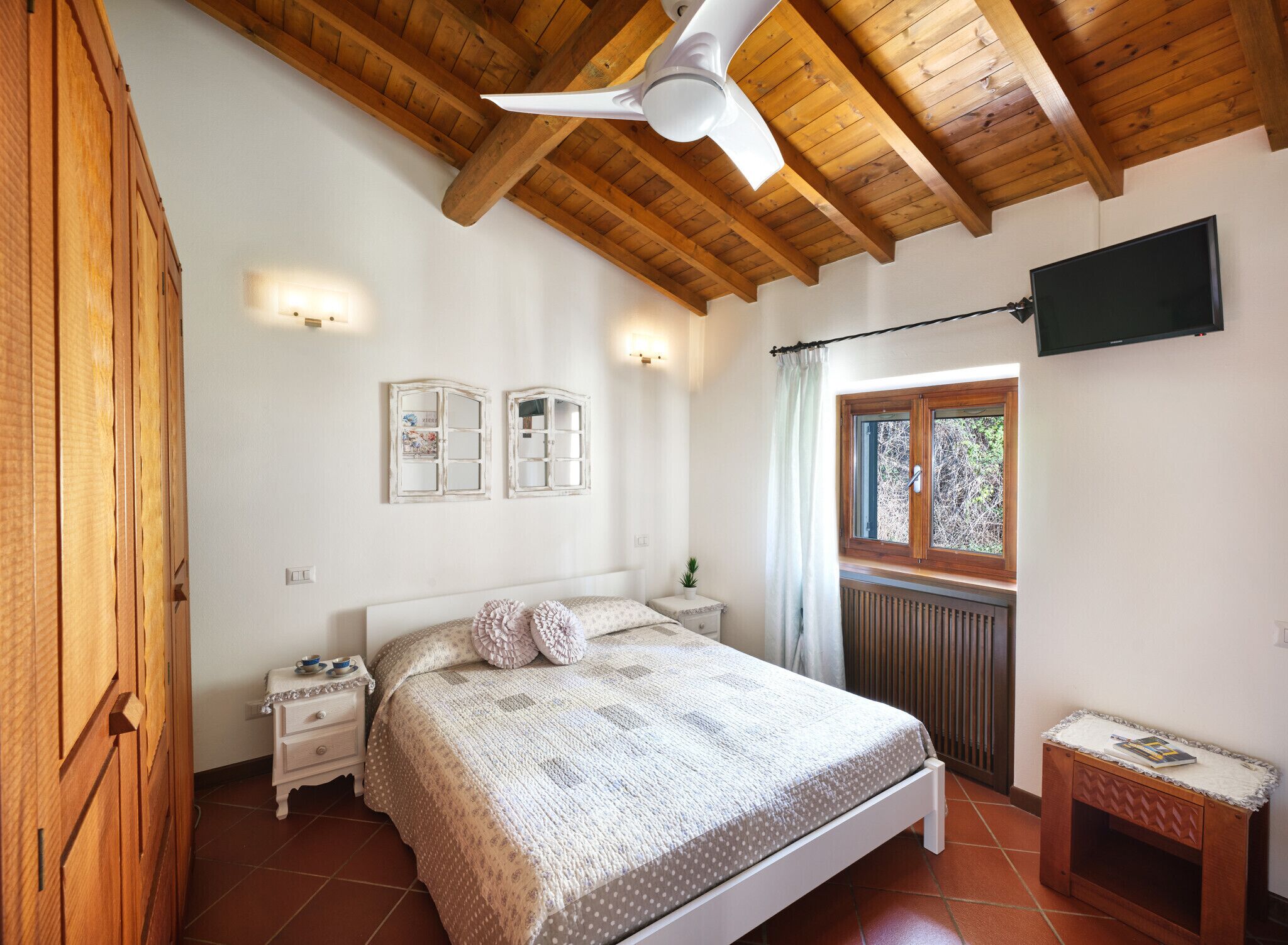 A bedroom with a wooden ceiling, a bed with a patterned bedspread, two wall-mounted light fixtures, a window with a view of greenery, and a small bedside table with a lamp.