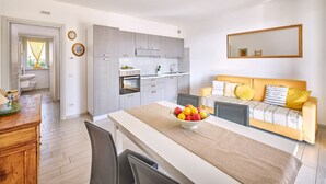 A modern kitchen with grey cabinets, a built-in oven, and a sink. There is a dining table with a fruit bowl and chairs. A yellow sofa with patterned cushions is against the wall.