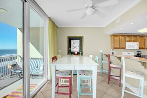 Un comedor con una mesa blanca y sillas de colores, una cocina con armarios de madera y vista al océano a través de una puerta corrediza de vidrio.