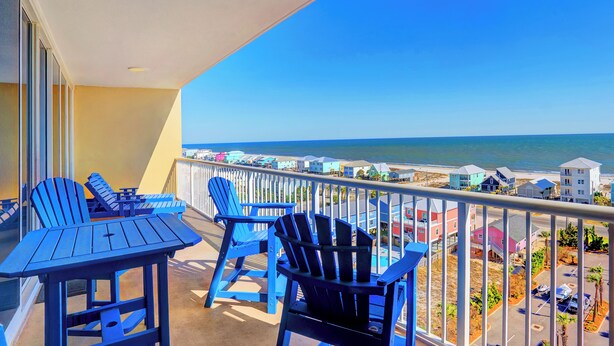 Ein Balkon mit blauen Adirondack-Stühlen und einem Tisch, mit Blick auf einen Strand mit bunten Gebäuden und dem Meer.