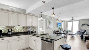 A modern kitchen with white cabinets, a black countertop, and stainless steel appliances. There is a dining area with a table and chairs, and a living room with a sofa and a coffee table. - Shoalwater 802 by Liquid Life (Orange Beach)