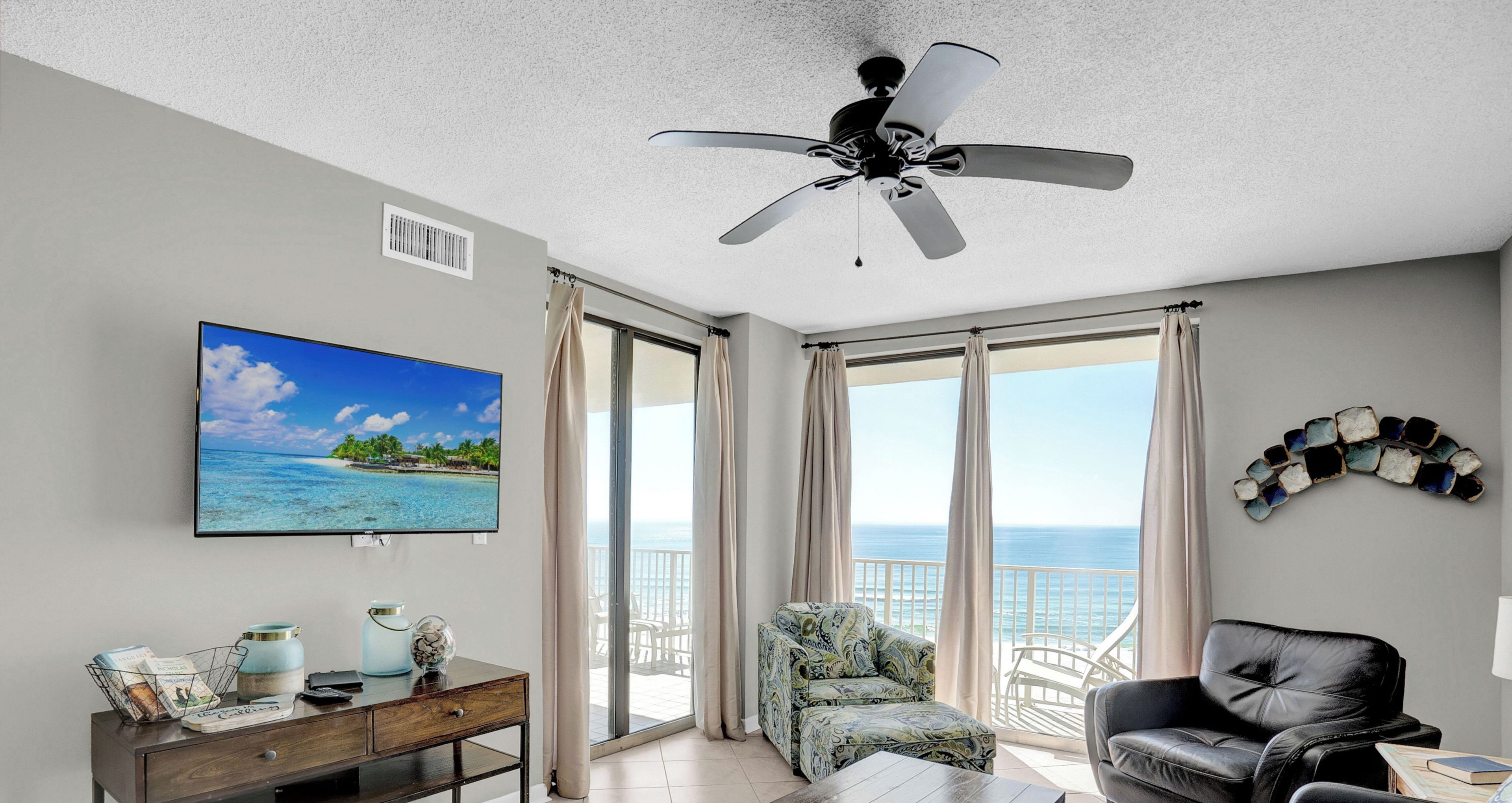 A living room with a ceiling fan, a television displaying a beach scene, a sofa, a coffee table, and a view of the ocean through large windows.