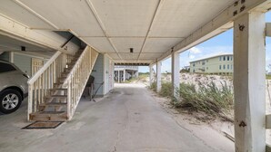 Ein überdachter Gang mit einer Treppe, die zu einem Gebäude führt, ein geparktes Auto und im Hintergrund eine Strand- und Wohngegend.