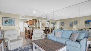 A living room with a light blue sofa, a white armchair, a wooden coffee table, and a dining area with wooden chairs and a table. - Surfside Shores 1302 by Liquid Life (Gulf Shores)