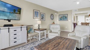 A living room with a flat-screen TV, white furniture, a coffee table, and a patterned rug. - Surfside Shores 1302 by Liquid Life (Gulf Shores)