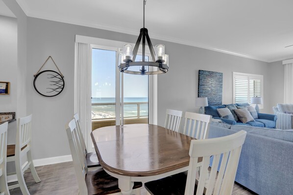 A dining area with a wooden table and white chairs, a chandelier, and a view of the ocean through a large window. - The Enclave 901 by Liquid Life (Orange Beach)