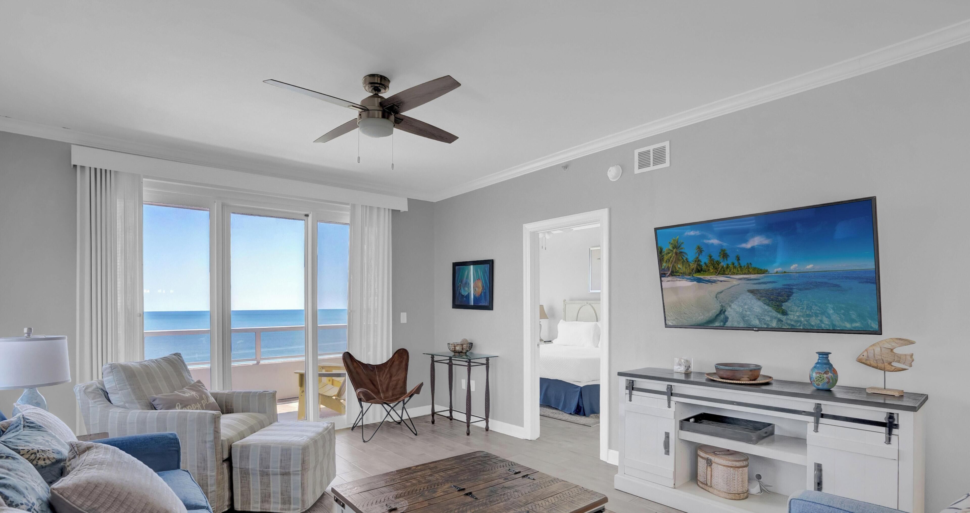 Ein Wohnzimmer mit Meerblick, einem Deckenventilator, einem großen Gemälde und einem Essbereich mit Tisch und Stühlen.