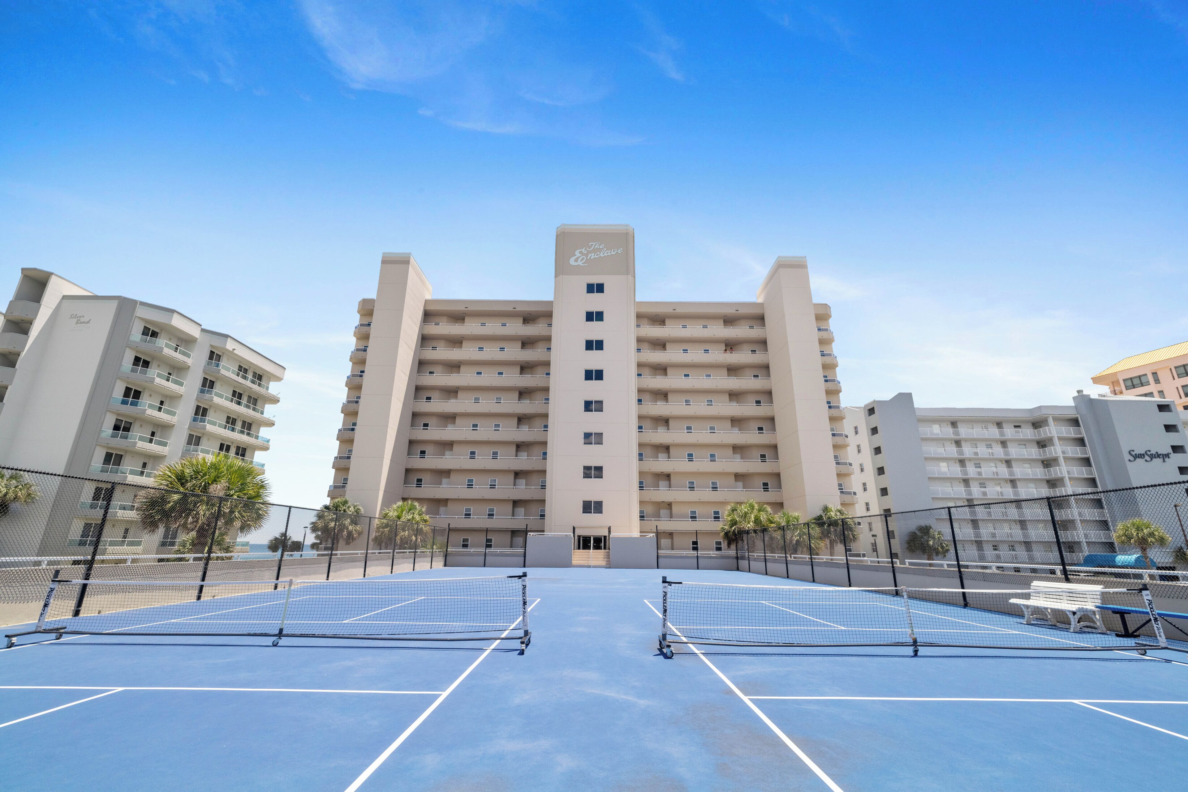 A tennis court with a net, surrounded by a fence, and buildings in the background.