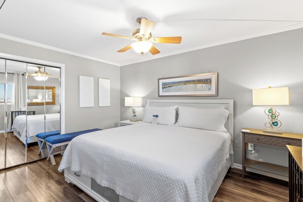 A hotel room with a large bed, a ceiling fan, a bedside table with a lamp, a framed picture on the wall, and a mirror on the door. - The Gulf Tower 3D by Liquid Life (Gulf Shores)