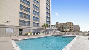A swimming pool with lounge chairs, a sign indicating pool rules, and a building with multiple balconies in the background.