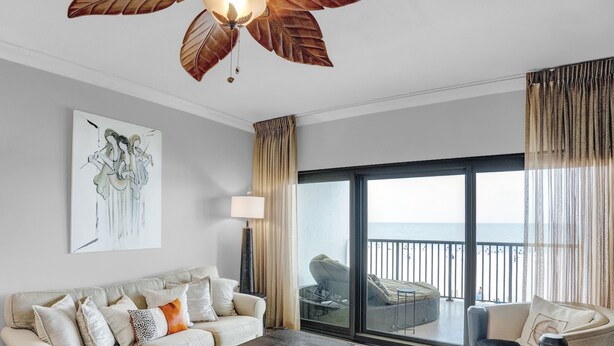 A modern living room with a large window, a ceiling fan, a sofa, and a painting on the wall. - The Palms 313 by Liquid Life (Orange Beach)