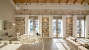 Un salón amplio con paredes de piedra, techo de madera, zona de comedor con mobiliario blanco y ventanas grandes con cortinas.