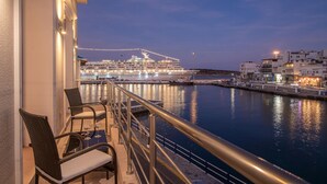 Un balcón con sillas de mimbre y una mesa, con vista a un puerto tranquilo donde un crucero está amarrado al fondo.