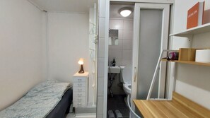 A small, compact room with a bed, a bedside table with a lamp, a wooden shelf with toiletries, and a view into the bathroom. - Only for woman - Sadang House (Seoul)