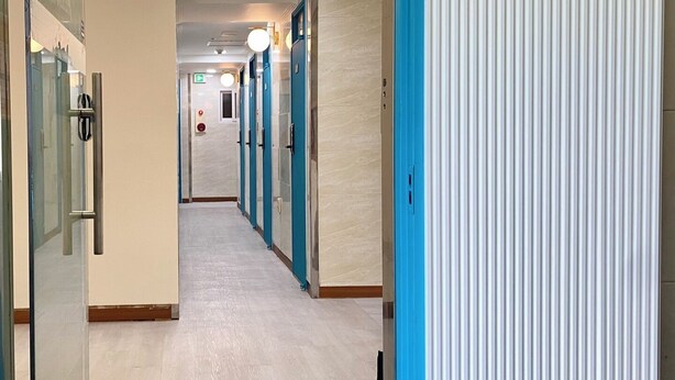 A hotel corridor with blue doors, a glass wall on the left, and a white wall on the right. - Only for woman - Sadang House (Seoul)