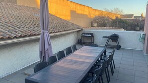 A rooftop patio with a long wooden table and chairs, a grill, and an umbrella.