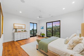 Ein Schlafzimmer mit Bett, Kommode, Stuhl und Blick ins Freie durch große Fenster.