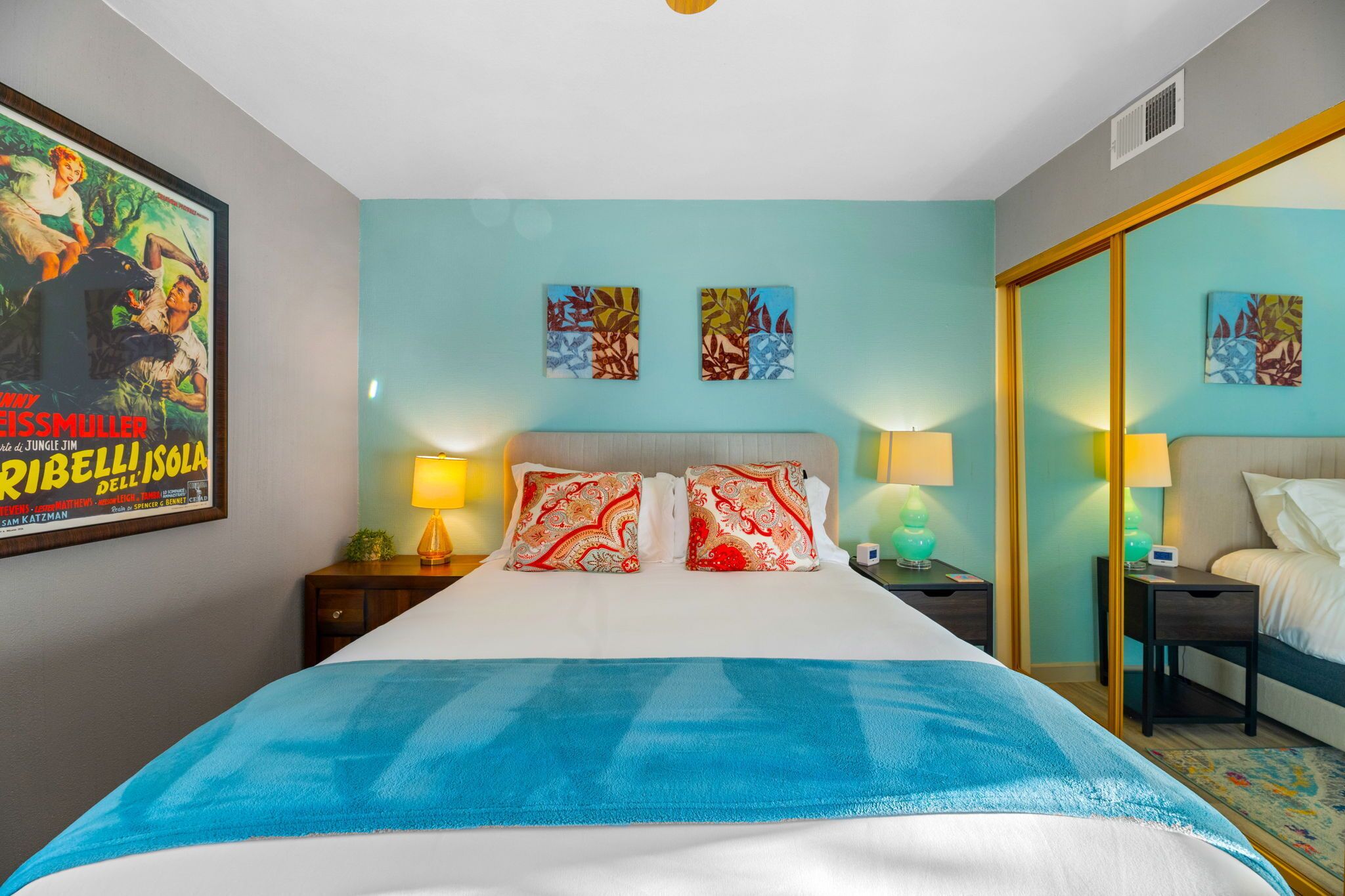 Une chambre d’hôtel avec un grand lit, deux tables de chevet avec des lampes, une petite table d’appoint, une affiche au mur et deux œuvres d’art décoratif.