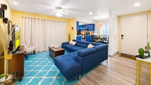 A living room with a sectional sofa, a television, a coffee table, and a dining area with blue cabinets and a kitchen in the background. - Beautiful Community! Pool, Jacuzzi, Tennis, Golf (Palm Springs)