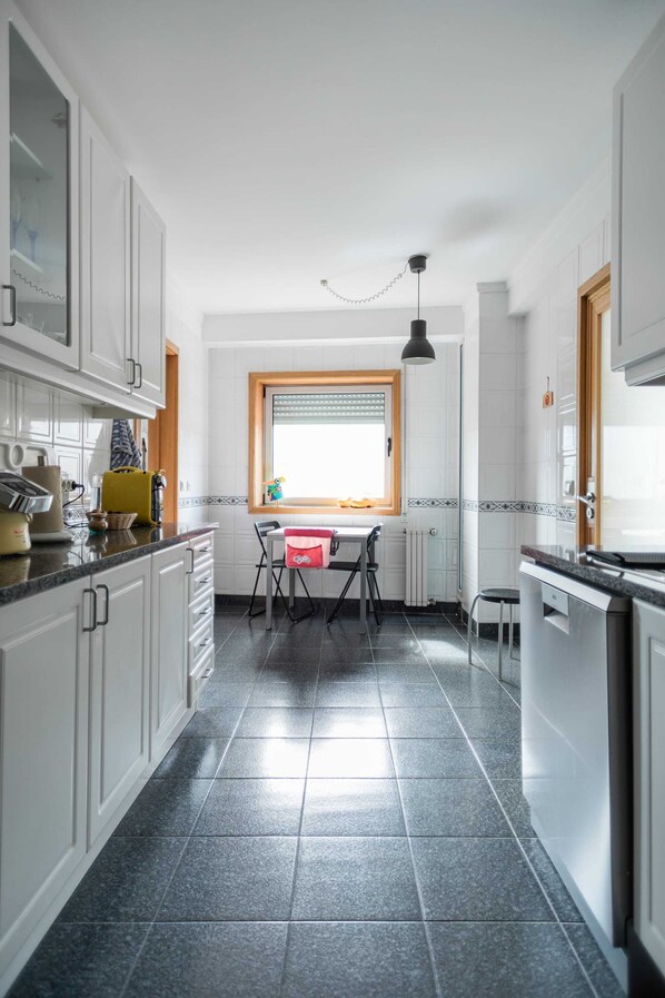 A kitchen with white cabinets, a dining table with chairs, a window with blinds, and a pendant light. - Gaia Beach&Golf – EcoHost | Parking · Surf · 5 pax (Arcozelo)