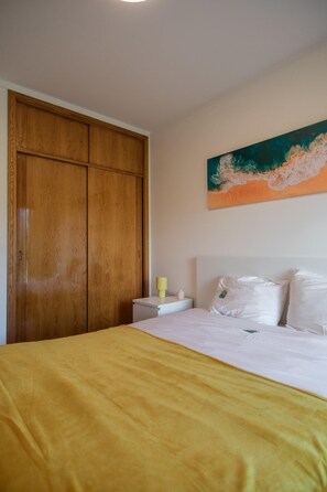 A bedroom with a bed, a wooden wardrobe, a nightstand with a lamp, and a painting on the wall. - Gaia Beach&Golf – EcoHost | Parking · Surf · 5 pax (Arcozelo)