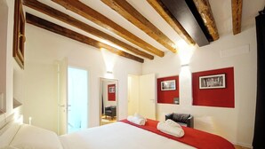 Ett hotellrum med en stor säng, synliga träbjälkar i taket, en röd accentvägg och en dörr som leder till ett annat rum.