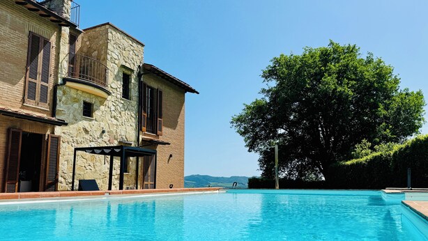 Una piscina junto a un edificio de piedra con contraventanas de madera y una pérgola.