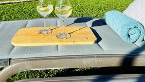 Una silla de lounge con una bandeja de madera que sostiene dos copas de vino blanco, frente a un césped verde exuberante y montañas distantes bajo un cielo azul despejado.