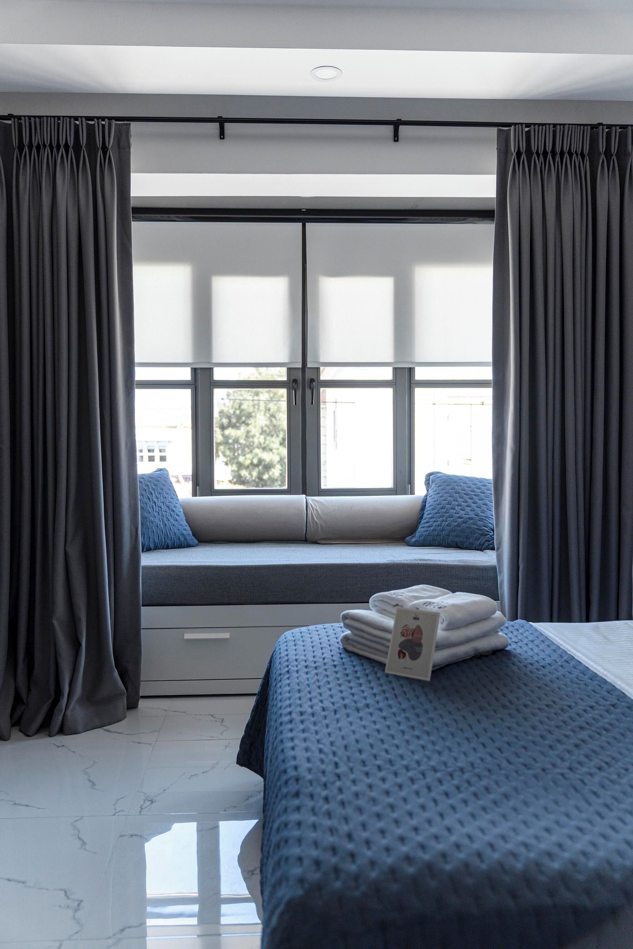 Moderni hotellihuone, jossa on suuri ikkuna, sisäänrakennettu penkki tyynyineen, siististi pedattu sänky ja pieni pöytä, jossa on pyyhkeitä.