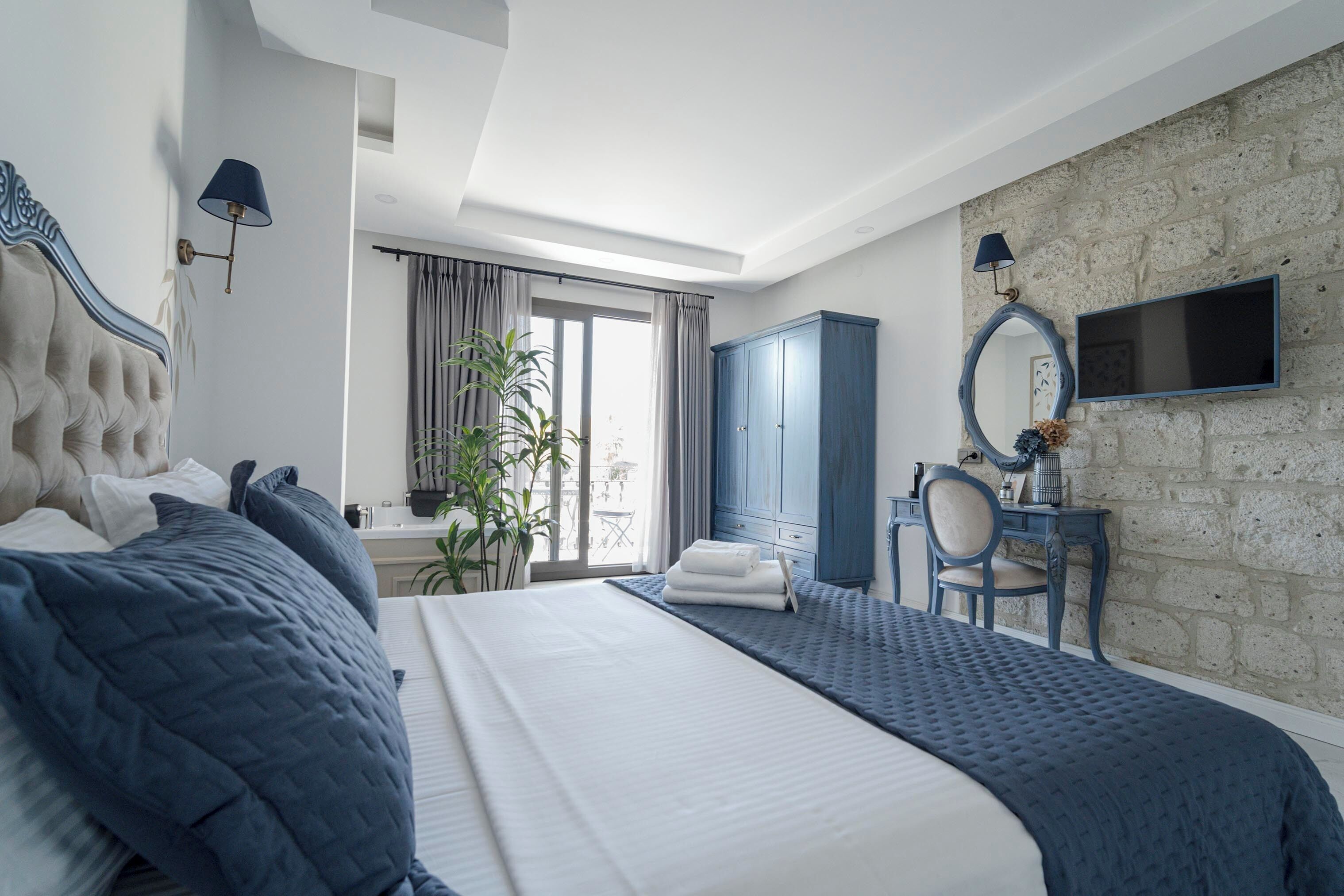 A modern bedroom with a large bed, a desk, a chair, a wardrobe, and a TV mounted on a stone wall.