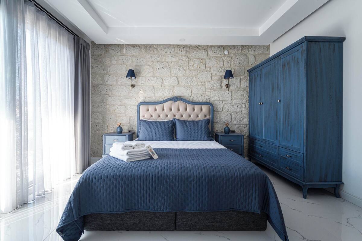 Ein Schlafzimmer mit Steinwand, einem gepolsterten Kopfteil, einer großen, blauen Tagesdecke, zwei Nachttischen und einem blauen Kleiderschrank.