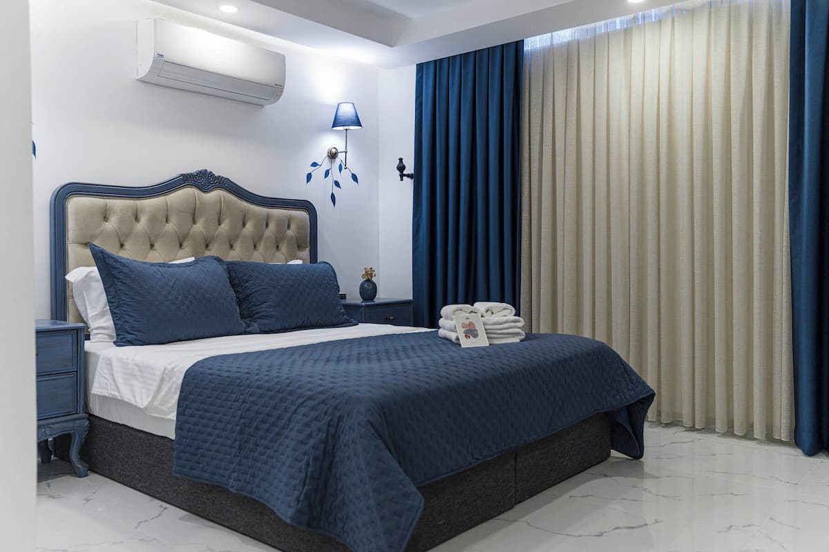 Ein Schlafzimmer mit einer gesteppten Kopfteile, einem ordentlich bezogenen Bett mit blauer Bettwäsche, einem Nachttisch, einer Wandlampe und einem Fenster mit blau-weißen Vorhängen.