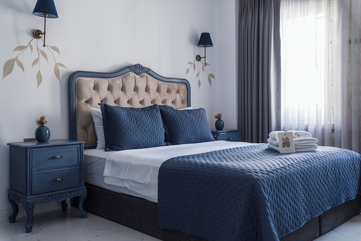 Ein Schlafzimmer mit einem gebürsteten Kopfteil, einer blauen gesteppten Bettdecke und passenden blauen Kissen. An jeder Seite des Bettes steht ein Nachttisch mit einer Vase und einer Lampe.