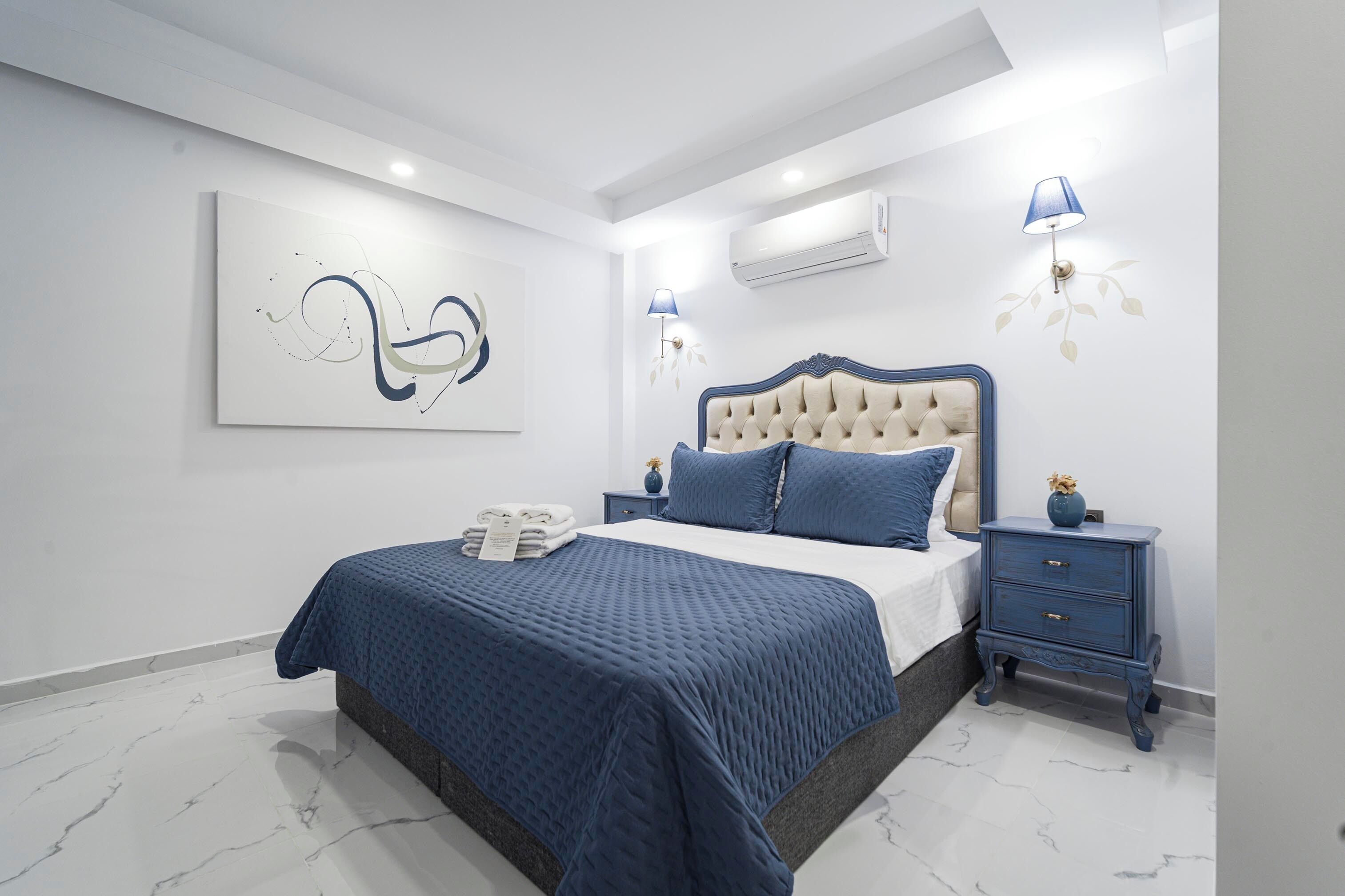 A modern bedroom with a large bed, a bedside table, a wall-mounted air conditioner, and a decorative wall piece.