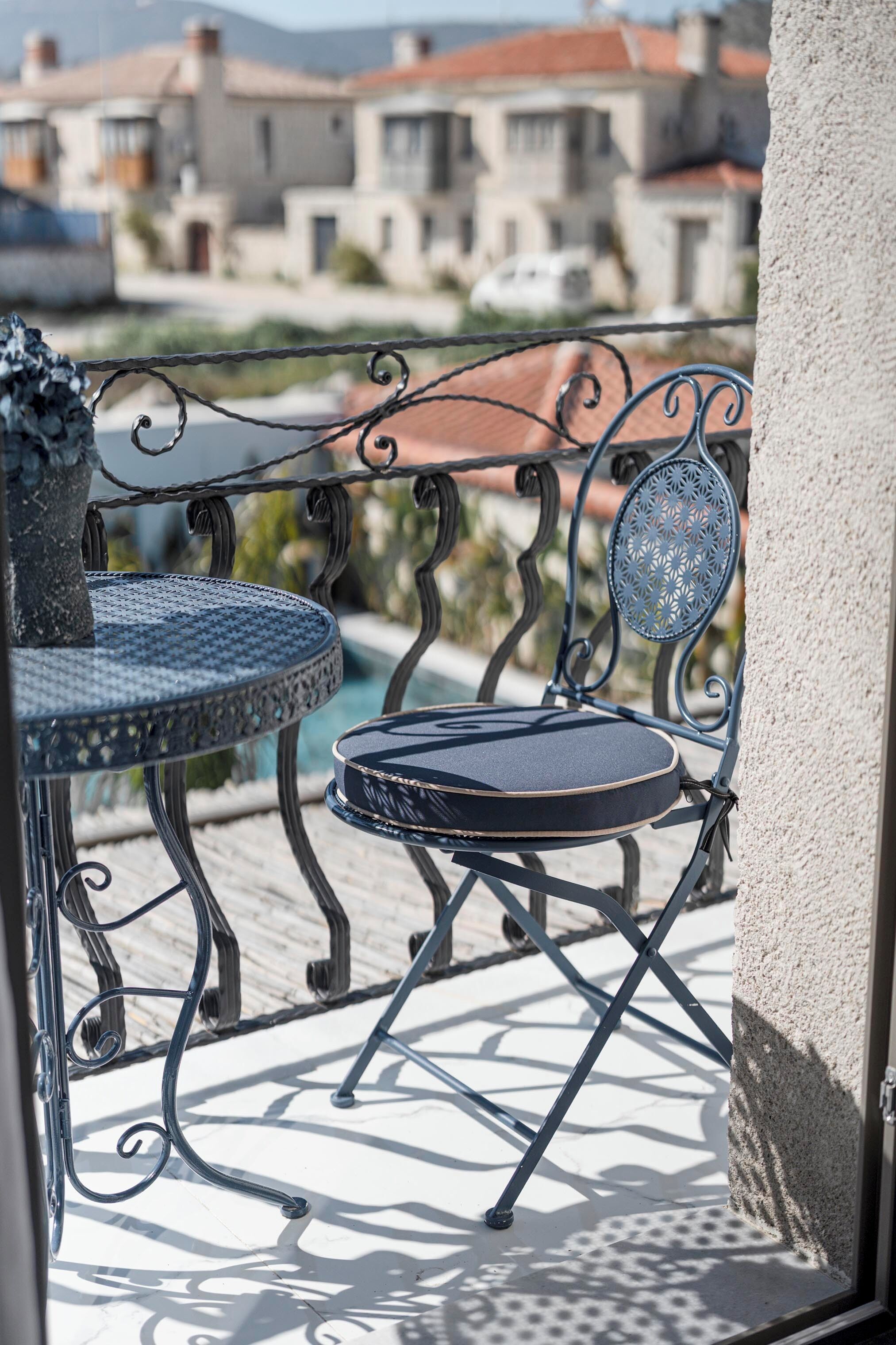 A balcony with a metal table and chair, a potted plant, and a view of buildings in the background.