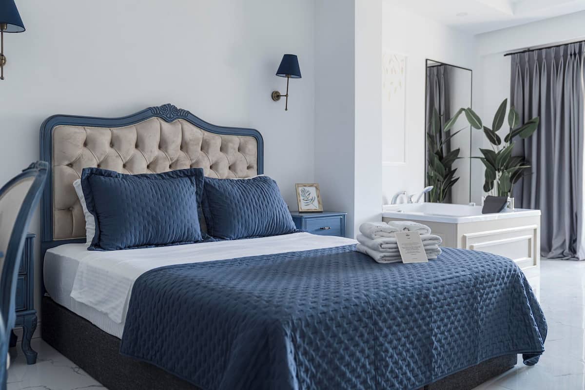 Ein Schlafzimmer mit einer gesteppten Kopfteil, einem großen Bett mit blauer Bettwäsche, einem Nachttisch mit Lampe, einem Spiegel und einem Badezimmer mit Waschbecken und Handtuchhalter.