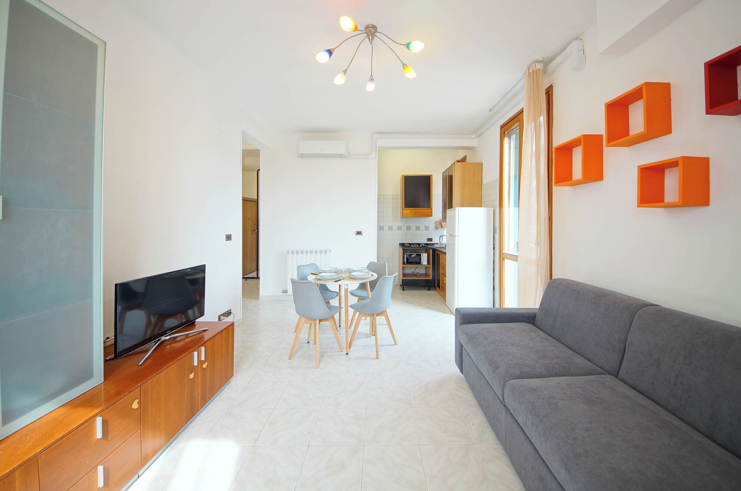 A modern living room with a flat-screen TV, grey sofa, dining table with chairs, and a kitchen area visible through an open doorway.