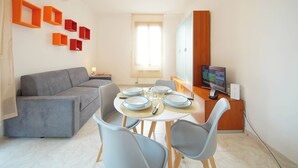 Ein modernes Wohnzimmer mit einem grauen Sofa, einem Esstisch gedeckt mit Tellern und Gläsern sowie einem Fernseher auf einer Holzkommode.